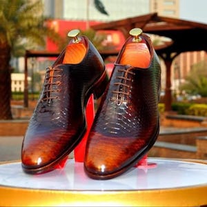 May include: A pair of brown leather oxfords with a snakeskin pattern. The shoes have lace-up closures and are on display on a round table.