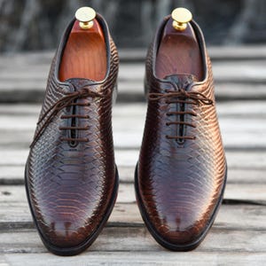 May include: A pair of brown leather oxfords with a snakeskin pattern. The shoes have lace-up closures and are on wooden shoe trees.
