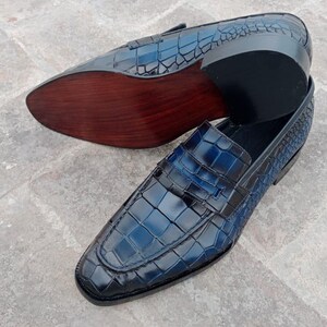Puede incluir: Un par de mocasines de cuero azul con estampado de cocodrilo. Los mocasines tienen una suela de cuero marr&oacute;n y una parte superior de cuero negro.