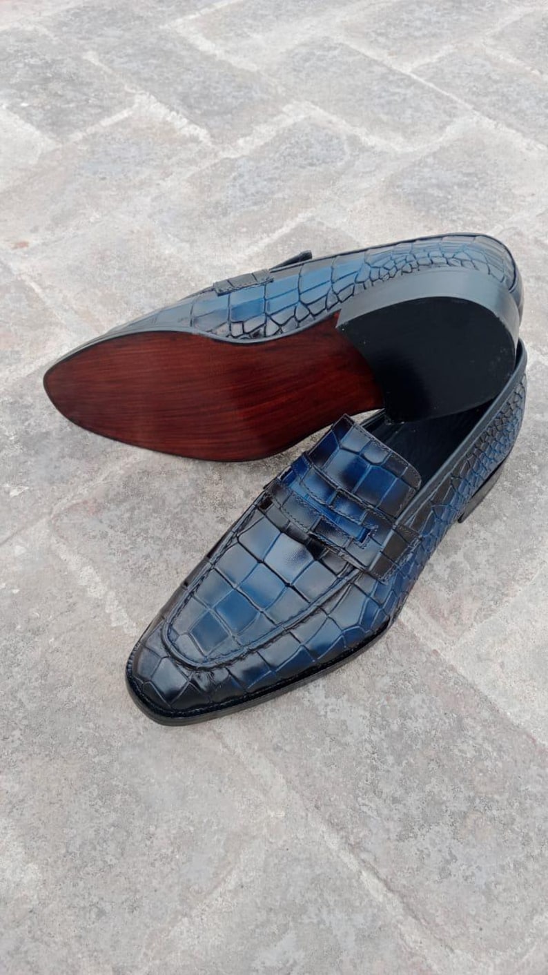Puede incluir: Un par de mocasines de cuero azul con estampado de cocodrilo. Los mocasines tienen una suela de cuero marr&oacute;n y una parte superior de cuero negro.