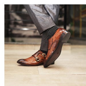 May include: Brown leather double monk strap shoes with a crocodile texture. The shoes have gold-colored buckles and a dark brown sole. The shoes are worn with gray pants and black and white striped socks.