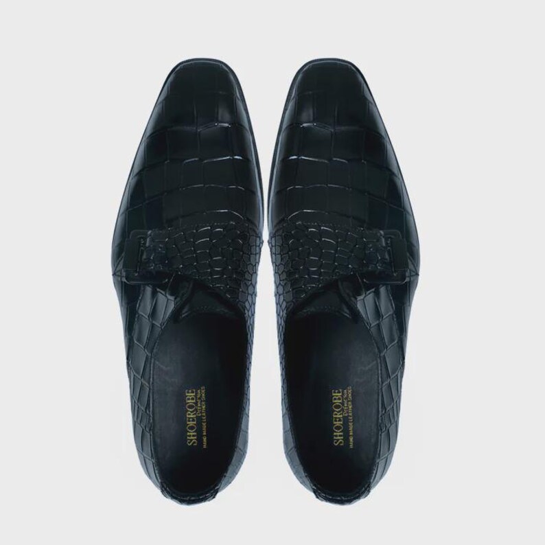 May include: A pair of black leather shoes with a crocodile pattern. The shoes have a buckle closure and a pointed toe.