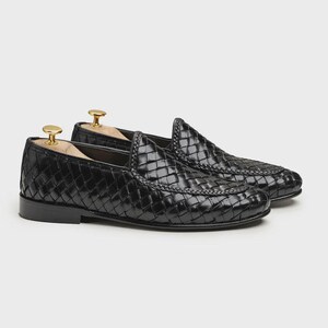 May include: A pair of black woven leather loafers with a classic design. The shoes have a glossy finish and a rounded toe. Shoe trees with gold-colored knobs are inserted in the shoes.