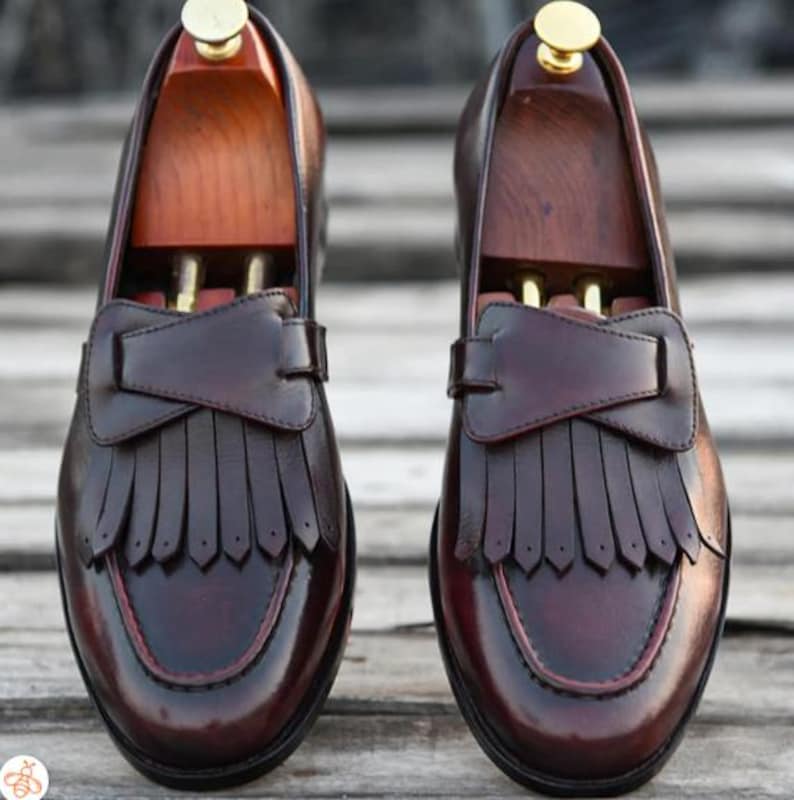 May include: A pair of brown leather loafers with fringe detailing. The loafers have a pointed toe and a low heel.