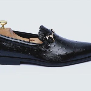 May include: Black leather loafer with a textured finish and a decorative buckle. The shoe features a classic design with a rounded toe and a low heel. A wooden shoe tree is inside the shoe.