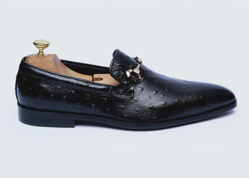 May include: Black leather loafer with a textured finish and a decorative buckle. The shoe features a classic design with a rounded toe and a low heel. A wooden shoe tree is inside the shoe.