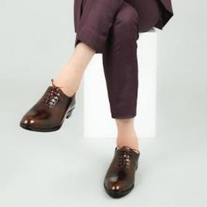 May include: A pair of brown leather lace-up shoes with a polished finish. The shoes are worn by a person sitting on a white stool, wearing burgundy pants.