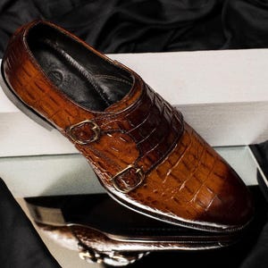 May include: A brown leather double monk strap shoe with a textured, crocodile-like pattern. The shoe features two gold-colored buckles and a polished finish, reflecting light. The shoe is set against a dark background.