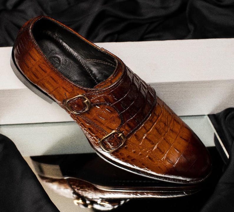 May include: A brown leather double monk strap shoe with a textured, crocodile-like pattern. The shoe features two gold-colored buckles and a polished finish, reflecting light. The shoe is set against a dark background.