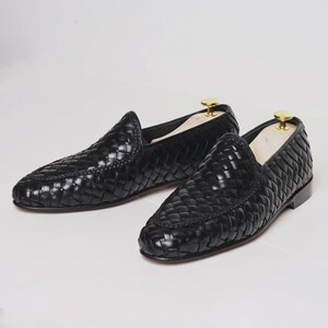 May include: A pair of black woven leather loafers. The shoes have a classic slip-on design with a rounded toe and a low heel. The leather has a glossy finish, and the shoes are displayed on a white surface.