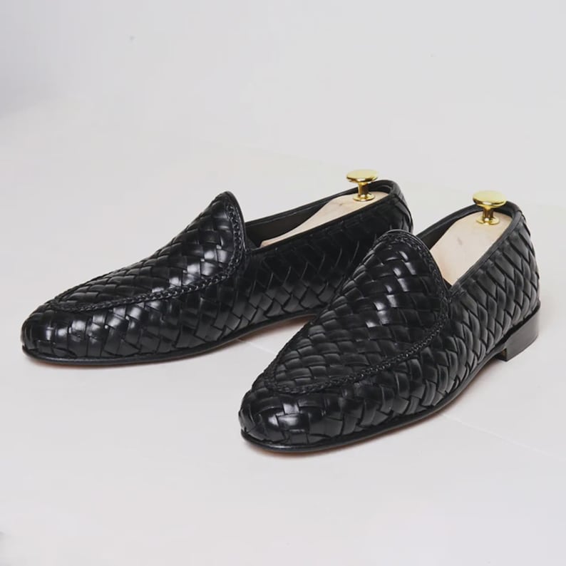 May include: A pair of black woven leather loafers. The shoes have a classic slip-on design with a rounded toe and a low heel. The leather has a glossy finish, and the shoes are displayed on a white surface.