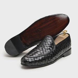 May include: Black woven leather loafers with a pointed toe. The sole is dark brown with a black heel. One shoe is upright, and the other is upside down, displaying the sole. A wooden shoe tree is inside the upright shoe.