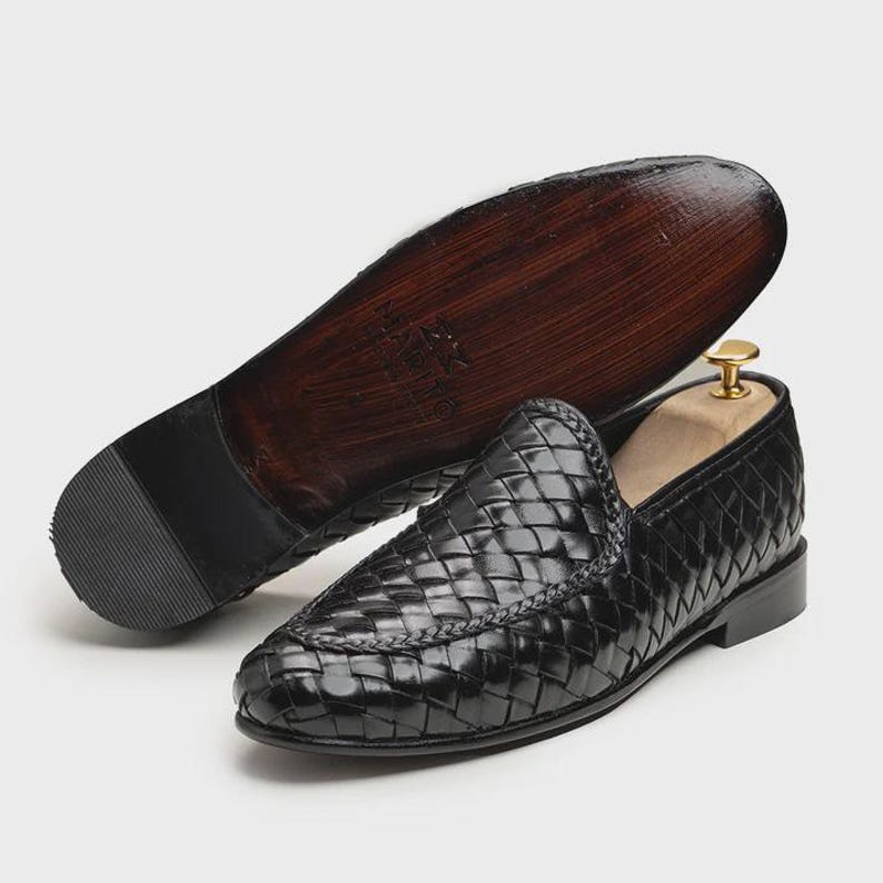 May include: Black woven leather loafers with a pointed toe. The sole is dark brown with a black heel. One shoe is upright, and the other is upside down, displaying the sole. A wooden shoe tree is inside the upright shoe.