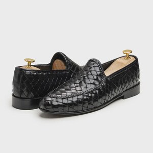 May include: A pair of black woven leather loafers. The shoes have a slip-on design with a rounded toe and a low heel. Shoe trees are inserted in the shoes. The leather has a glossy finish.