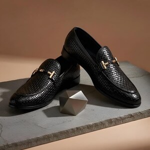 May include: A pair of black woven leather loafers with gold-colored metal accents. The shoes are displayed on a gray surface with a geometric silver-colored object in the foreground. The loafers have a classic, slip-on design.