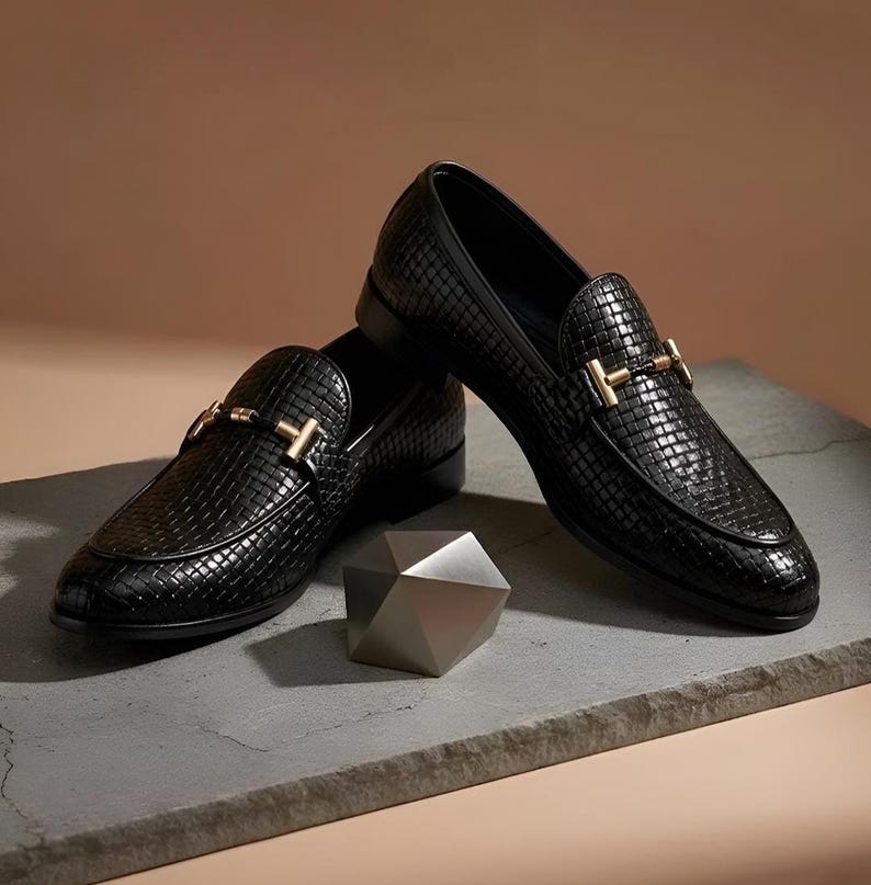 May include: A pair of black woven leather loafers with gold-colored metal accents. The shoes are displayed on a gray surface with a geometric silver-colored object in the foreground. The loafers have a classic, slip-on design.