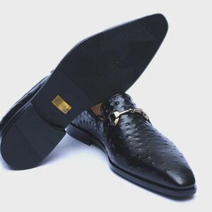 May include: Black leather slip-on dress shoes with a textured pattern and a gold-colored buckle. The sole of one shoe is visible, showing a textured pattern. The shoes are displayed on a white background.