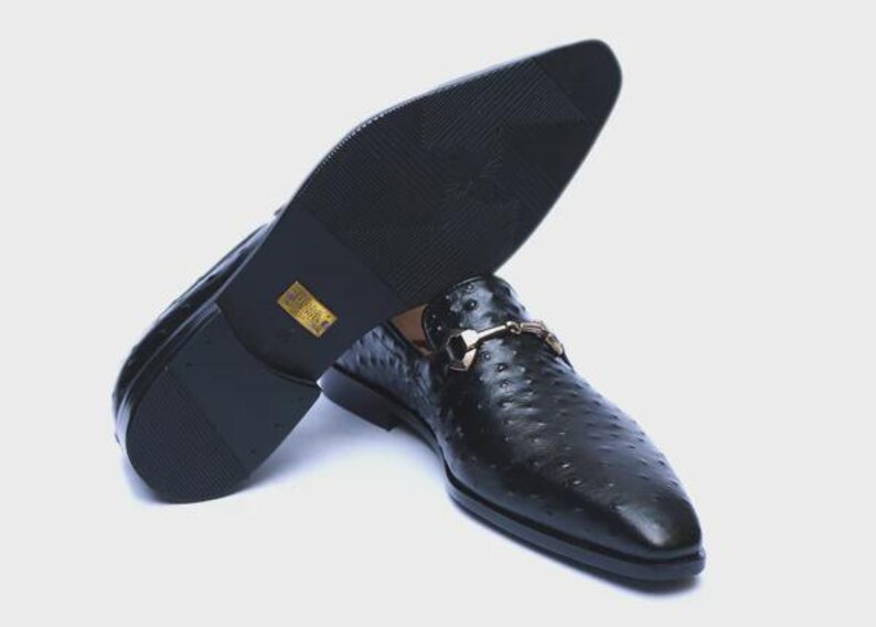 May include: Black leather slip-on dress shoes with a textured pattern and a gold-colored buckle. The sole of one shoe is visible, showing a textured pattern. The shoes are displayed on a white background.