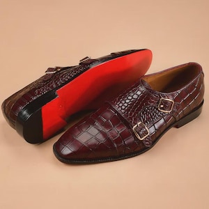 May include: A pair of burgundy leather double monk strap shoes with a crocodile pattern. The shoes have gold-colored buckles and a red sole. The shoes are displayed on a neutral background.