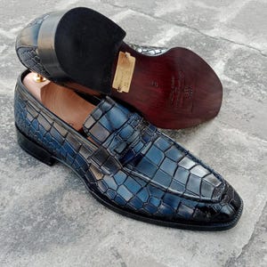 Puede incluir: Un par de mocasines azules de cuero con estampado de cocodrilo y detalle de hebilla. Los zapatos tienen una suela de cuero marr&oacute;n y un tac&oacute;n de cuero negro.