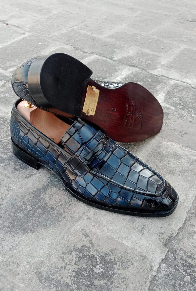 Puede incluir: Un par de mocasines azules de cuero con estampado de cocodrilo y detalle de hebilla. Los zapatos tienen una suela de cuero marr&oacute;n y un tac&oacute;n de cuero negro.