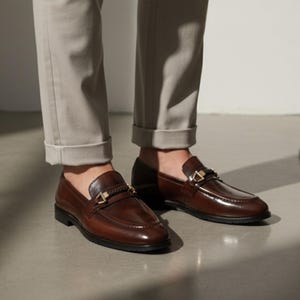 May include: Brown leather loafers with gold-colored accents. The shoes have a classic design with a polished finish and a decorative strap across the top. The loafers are worn with light-colored pants.