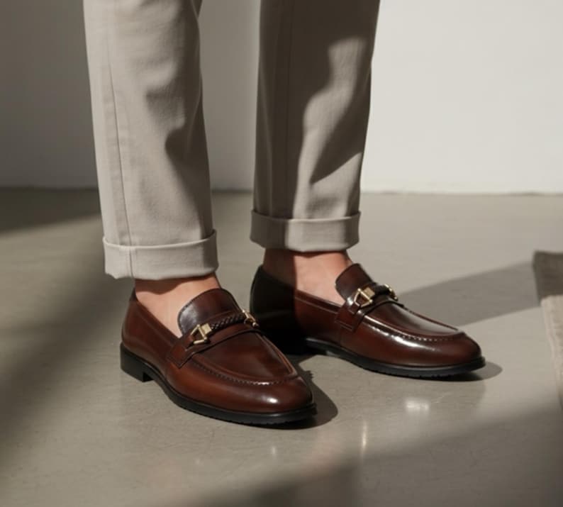 May include: Brown leather loafers with gold-colored accents. The shoes have a classic design with a polished finish and a decorative strap across the top. The loafers are worn with light-colored pants.