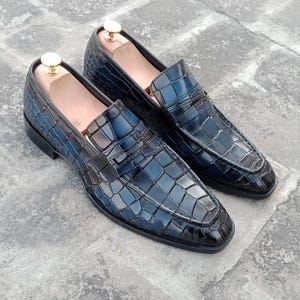 Puede incluir: Un par de mocasines de cuero negro con estampado de cocodrilo. Los mocasines tienen punta puntiaguda y un dise&ntilde;o de deslizamiento.
