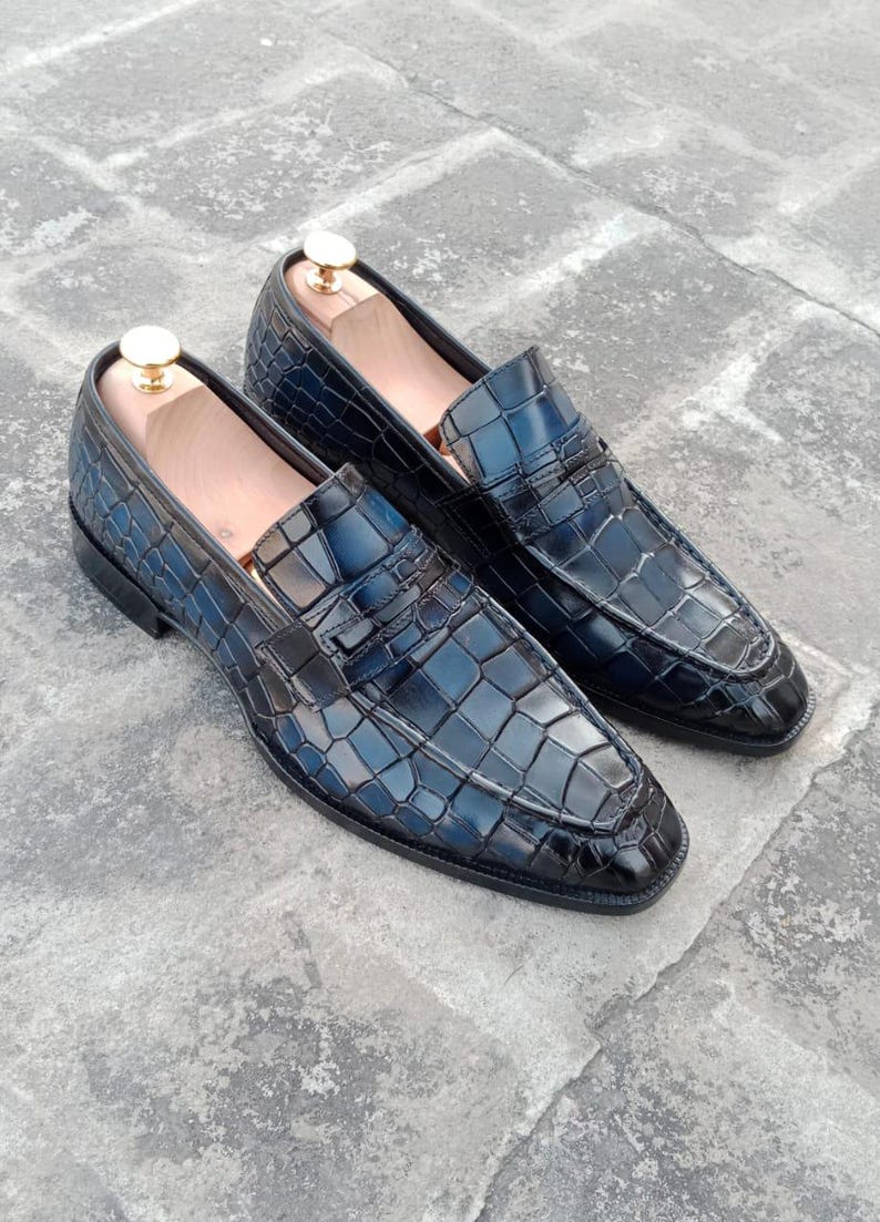 Puede incluir: Un par de mocasines de cuero negro con estampado de cocodrilo. Los mocasines tienen punta puntiaguda y un dise&ntilde;o de deslizamiento.