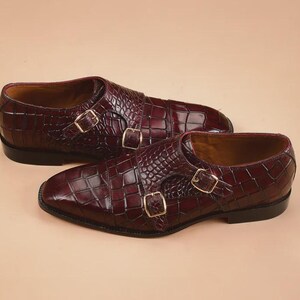 May include: A pair of burgundy leather double monk strap shoes with a crocodile skin texture. The shoes feature gold-colored buckles and a classic design, perfect for formal occasions.