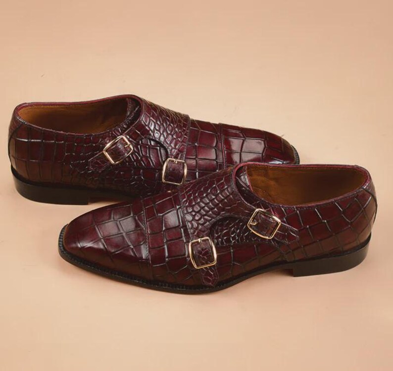 May include: A pair of burgundy leather double monk strap shoes with a crocodile skin texture. The shoes feature gold-colored buckles and a classic design, perfect for formal occasions.