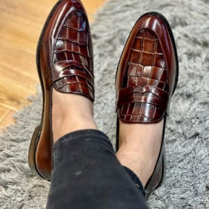 May include: A pair of brown leather loafers with a glossy finish. The shoes feature a textured, crocodile-like pattern on the vamp and a classic penny loafer design. The shoes are worn by a person wearing black pants.