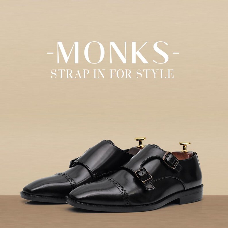 Puede incluir: Zapatos negros de cuero con hebilla de monje y doble cierre de hebilla. Los zapatos tienen un acabado pulido y se muestran sobre un fondo neutro. El texto "-MONKS- STRAP IN FOR STYLE" est&aacute; en la parte superior.