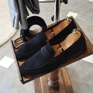 May include: A pair of black suede loafers with wooden shoe trees inside, displayed on a wooden tray. A black leather belt and a gray jacket are also visible. The shoes have a classic design.