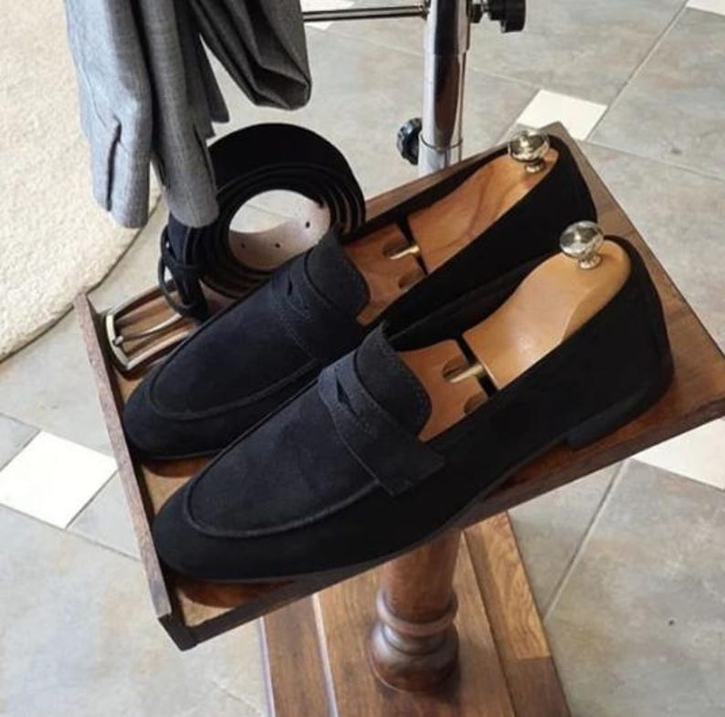 May include: A pair of black suede loafers with wooden shoe trees inside, displayed on a wooden tray. A black leather belt and a gray jacket are also visible. The shoes have a classic design.