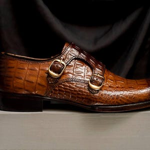 May include: Brown leather double monk strap shoe with a crocodile texture. The shoe features two gold-colored buckles and a dark brown sole. The shoe is displayed on a light gray surface against a black backdrop.