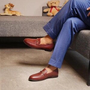 May include: Brown leather loafers with a gold-colored buckle detail. The shoes are a rich, reddish-brown color and have a classic, slip-on design. The loafers are worn with blue pants.