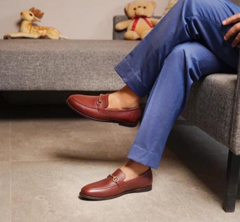 May include: Brown leather loafers with a gold-colored buckle detail. The shoes are a rich, reddish-brown color and have a classic, slip-on design. The loafers are worn with blue pants.