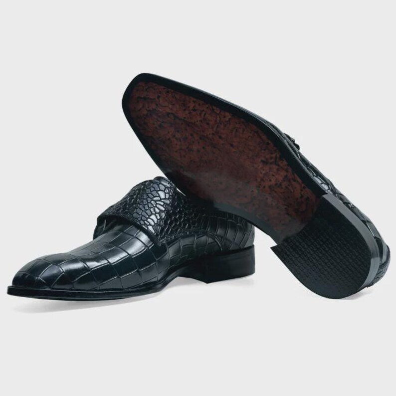 May include: A pair of black leather dress shoes with a crocodile pattern. The shoes have a strap across the top and a pointed toe.