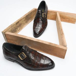 Op de afbeelding: Bruine leren herenschoenen met een krokodillenpatroon. De schoenen hebben een gespsluiting en worden gepresenteerd op een houten constructie tegen een witte achtergrond. Het leer heeft een glanzende afwerking.