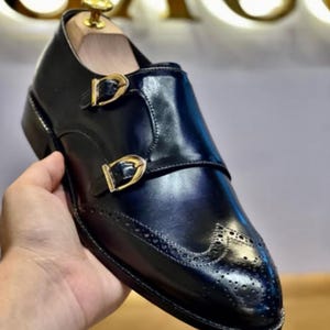 May include: Black leather double monk strap shoe with gold-coloured buckles. The shoe has decorative perforations on the toe and a polished finish. A wooden shoe tree is inserted inside the shoe.