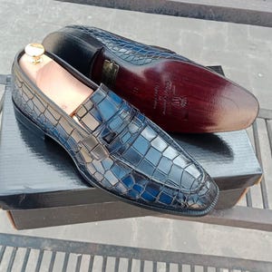 Puede incluir: Un par de mocasines azules de cuero con estampado de cocodrilo y suela de cuero marr&oacute;n. Los zapatos tienen una hebilla dorada y un forro de cuero negro. Los zapatos est&aacute;n sobre una caja negra con el nombre de la marca "Ciggy Couture" impreso en la suela.