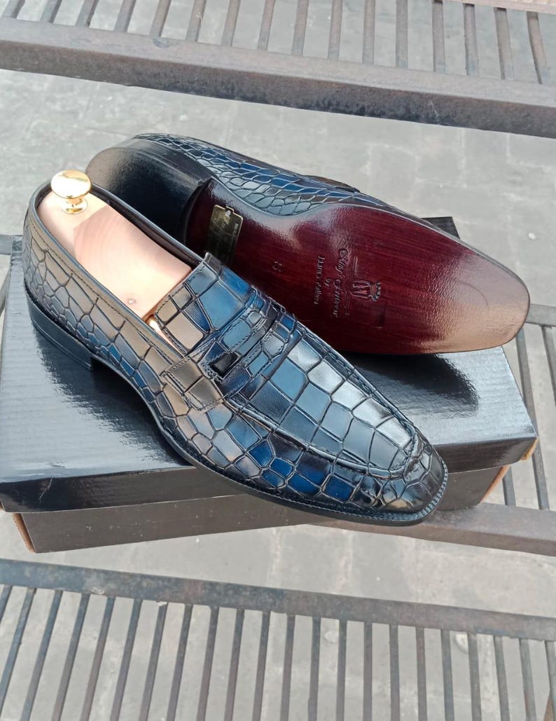 Puede incluir: Un par de mocasines azules de cuero con estampado de cocodrilo y suela de cuero marr&oacute;n. Los zapatos tienen una hebilla dorada y un forro de cuero negro. Los zapatos est&aacute;n sobre una caja negra con el nombre de la marca "Ciggy Couture" impreso en la suela.