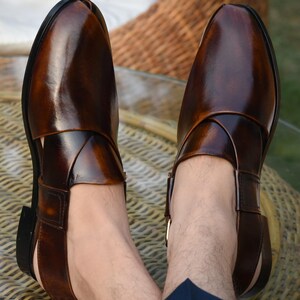 May include: A pair of brown leather sandals with a glossy finish. The sandals feature a unique design with straps crossing over the foot and a buckle closure. The open-toe design and dark brown color give a classic look.