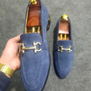 May include: A pair of blue suede loafers with gold-colored metal accents. The shoes have a classic slip-on design with a decorative buckle. The image shows the shoes on a neutral background.