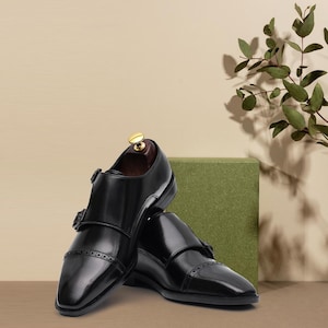 Puede incluir: Un par de zapatos de cuero negro brillante con doble hebilla y dise&ntilde;o de puntera. Los zapatos se exhiben sobre un fondo neutro, con un bloque verde y una ramita de vegetaci&oacute;n.