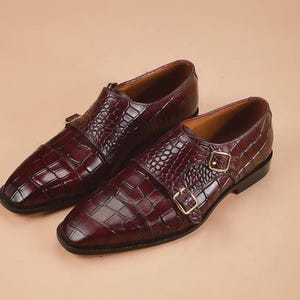 May include: A pair of burgundy leather double monk strap shoes. The shoes feature a textured, crocodile-like pattern and gold-colored buckles. The shoes have a polished finish and a classic design.