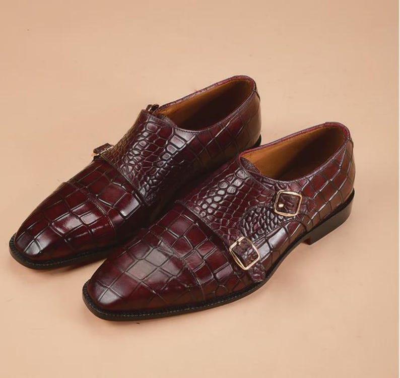 May include: A pair of burgundy leather double monk strap shoes. The shoes feature a textured, crocodile-like pattern and gold-colored buckles. The shoes have a polished finish and a classic design.