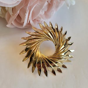 May include: A circular gold-tone brooch featuring a sunburst design with multiple radiating, pointed metal petals that spiral outward from a hollow center. The polished metallic surface reflects light, creating a bright, reflective finish.