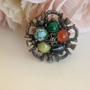 May include: A vintage brooch with a silver-tone metal thistle design. The brooch features six colorful, round stones in shades of blue, green, orange, yellow, and black. The brooch is a decorative accessory.